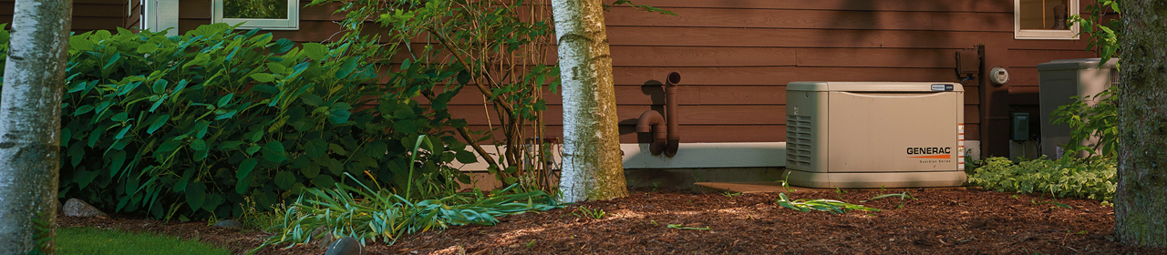 Generac Home Standby Generator behind a red house between trees and bushes.