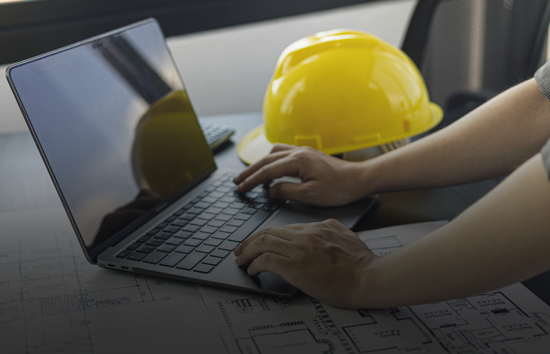 Contractor using a laptop with blueprints and hard hat nearby representing Generac's industrial generator TCO calculator to estimate diesel and natural gas generator costs