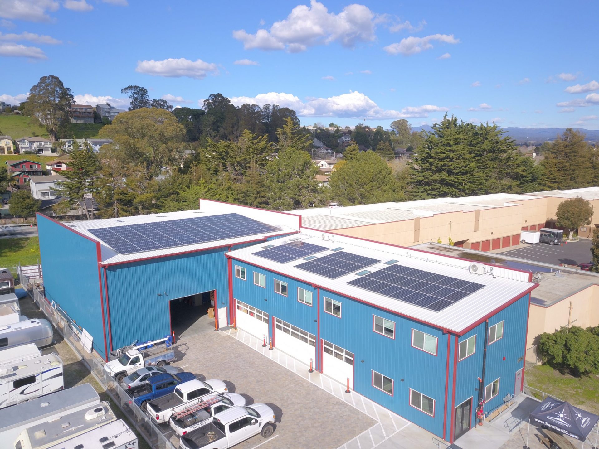 Aerial view of Sandbar Solar’s blue-roofed facility in Santa Cruz with solar panels installed across two rooftops, powered by a Generac microgrid