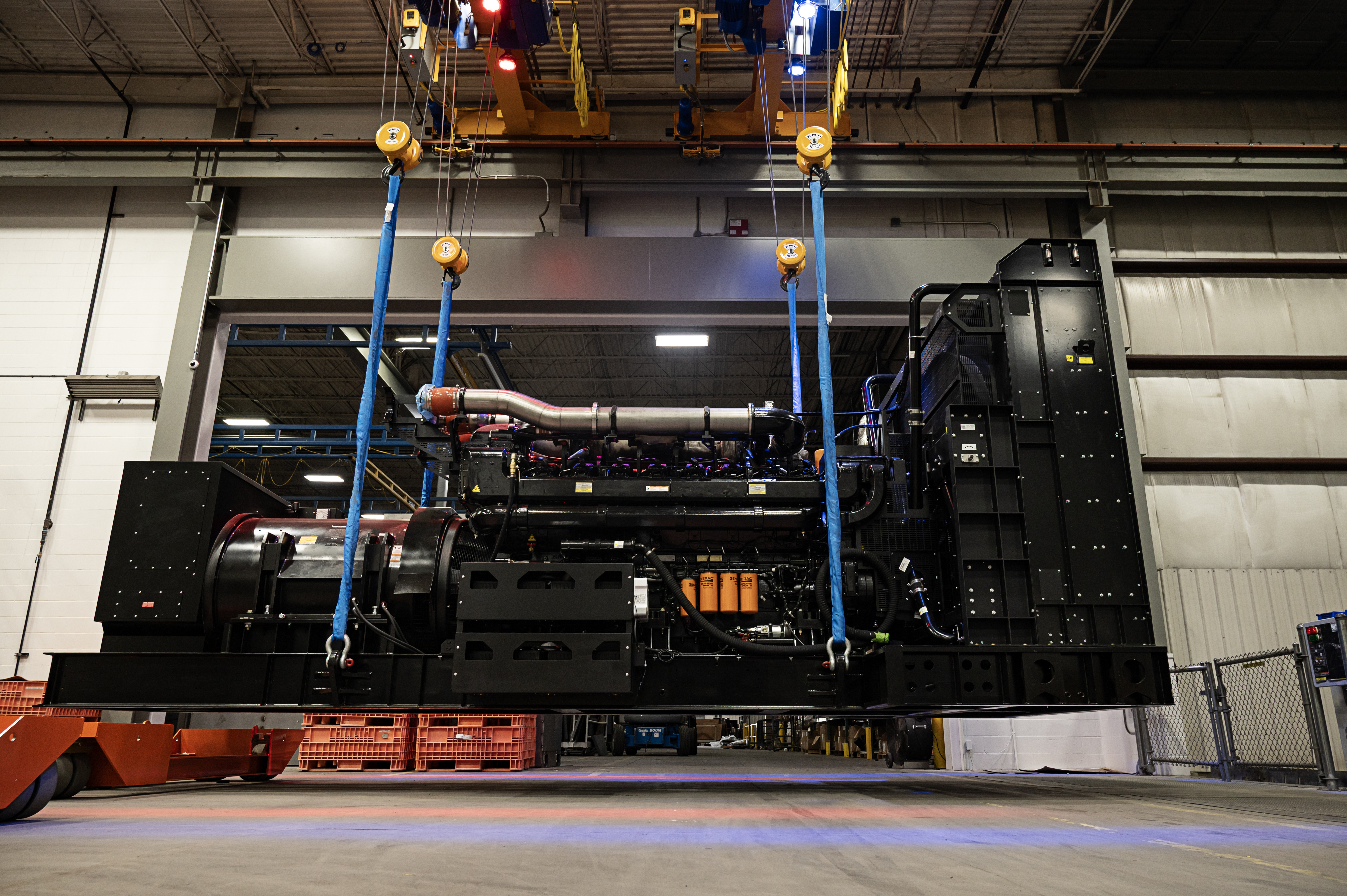 A large black generator is held by an overhead crane at a manfuacturing facility