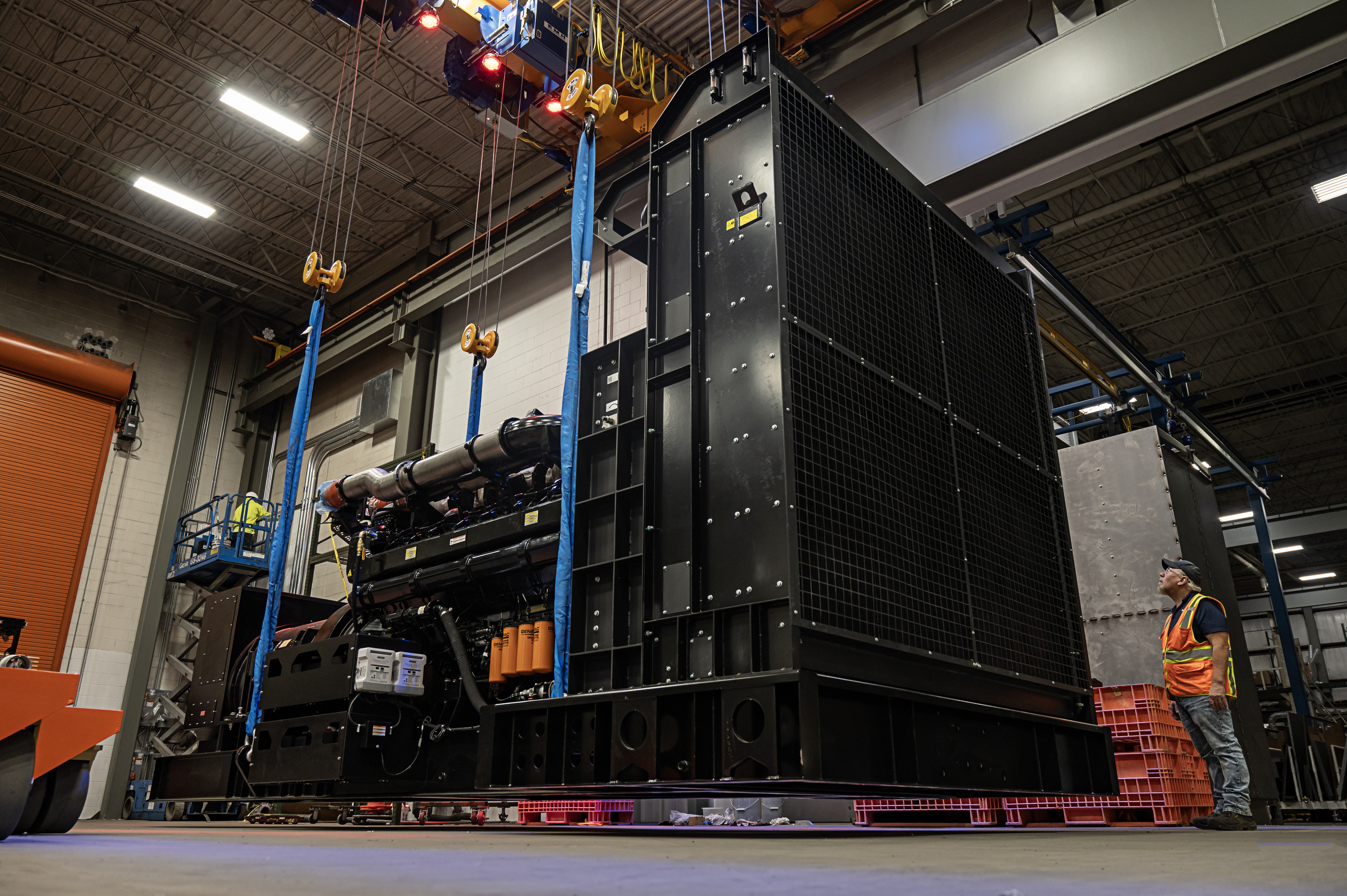  A man stands next to a large black generator being held in the air by an overhead crane