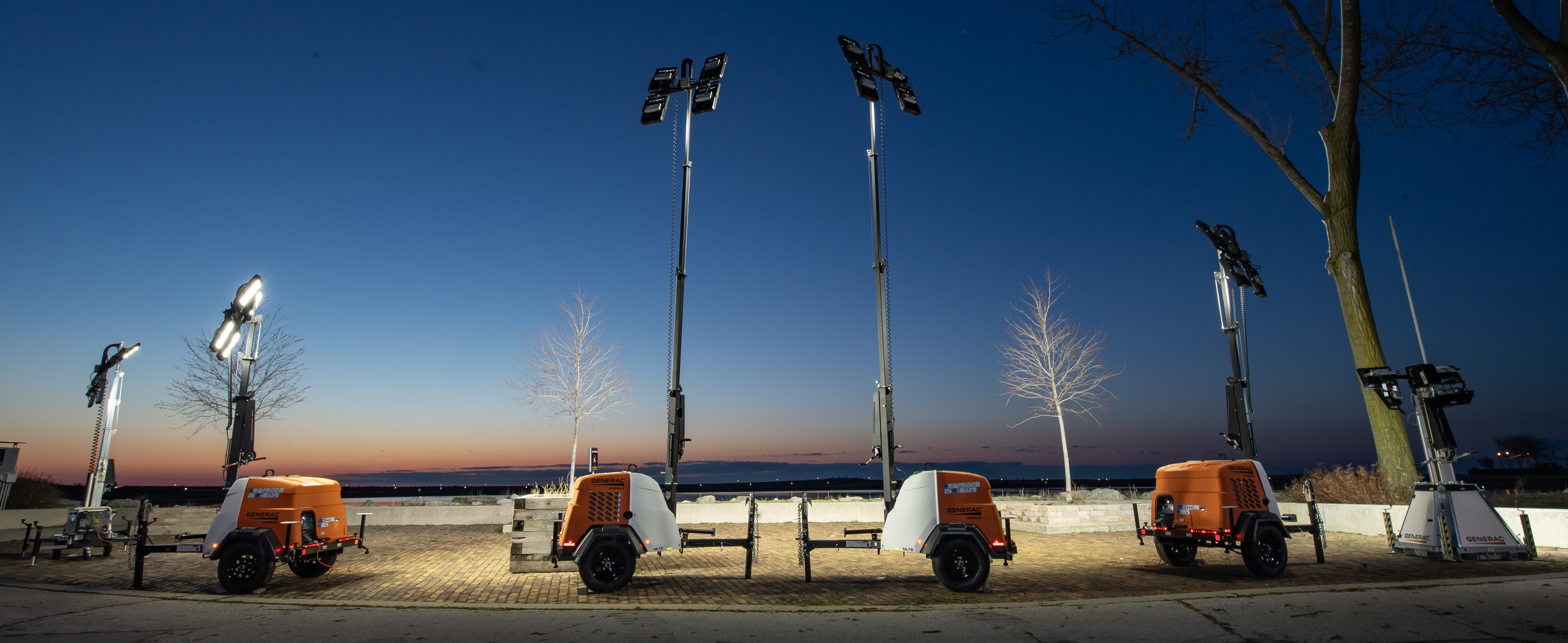  A series of orange, black and white Generac light towers site in a parking lot at sunset